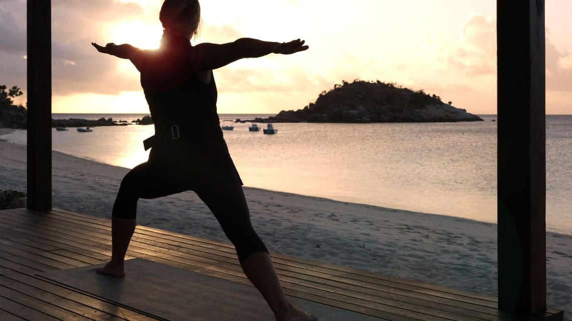 Person practicing yoga in a warrior pose on a wooden deck by the beach at sunset. The scene is tranquil, with an island and boats in the distance.