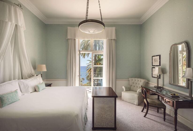Elegant hotel room with soft green walls, a large bed with a canopy, and a window view of the ocean and palm trees. A desk and armchair add a cozy touch.