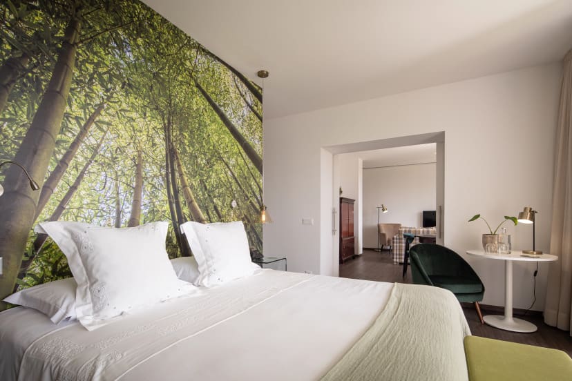 Modern hotel room with a nature-themed mural of tall green trees above the bed. Bright, airy space with elegant decor, including a small table and chair.