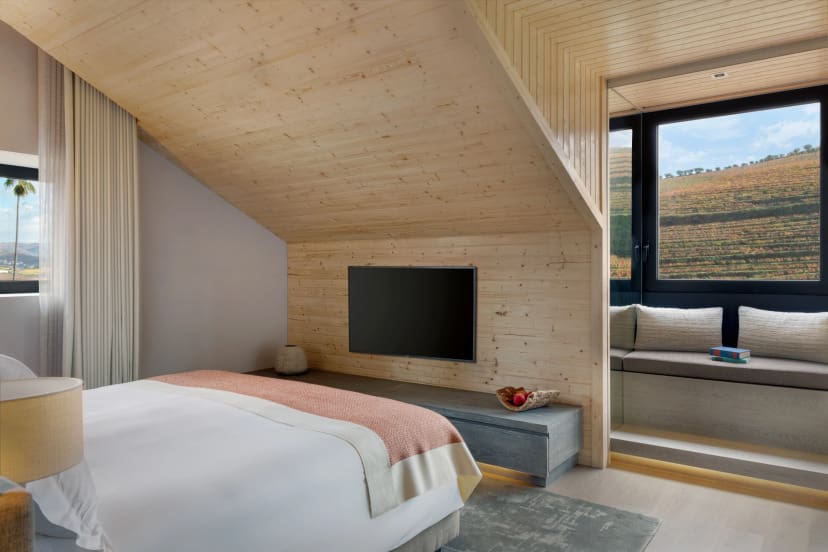 A cozy bedroom with a sloped wooden ceiling, a neatly made bed with beige and coral bedding, a wall-mounted TV, and a window seat offering a vineyard view.