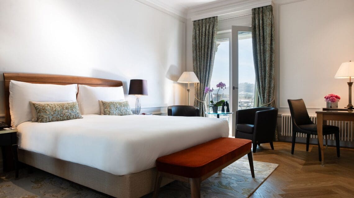 A cozy hotel room with a large bed and white linens, flanked by modern lamps. Two armchairs by a window with curtains, overlooking a scenic view.