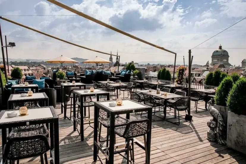 Rooftop terrace with wicker tables and chairs, shaded by umbrellas. Overlooks city skyline under a partly cloudy sky. Inviting and relaxed atmosphere.