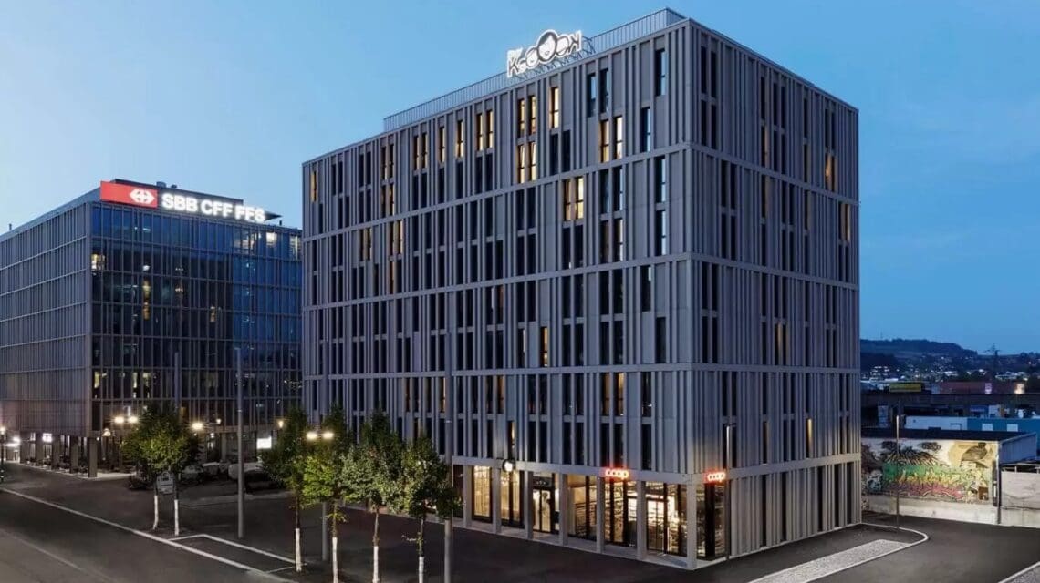 Modern multi-story building at dusk with vertical lines and large windows, flanked by another tall structure. Streetlights illuminate the area below.