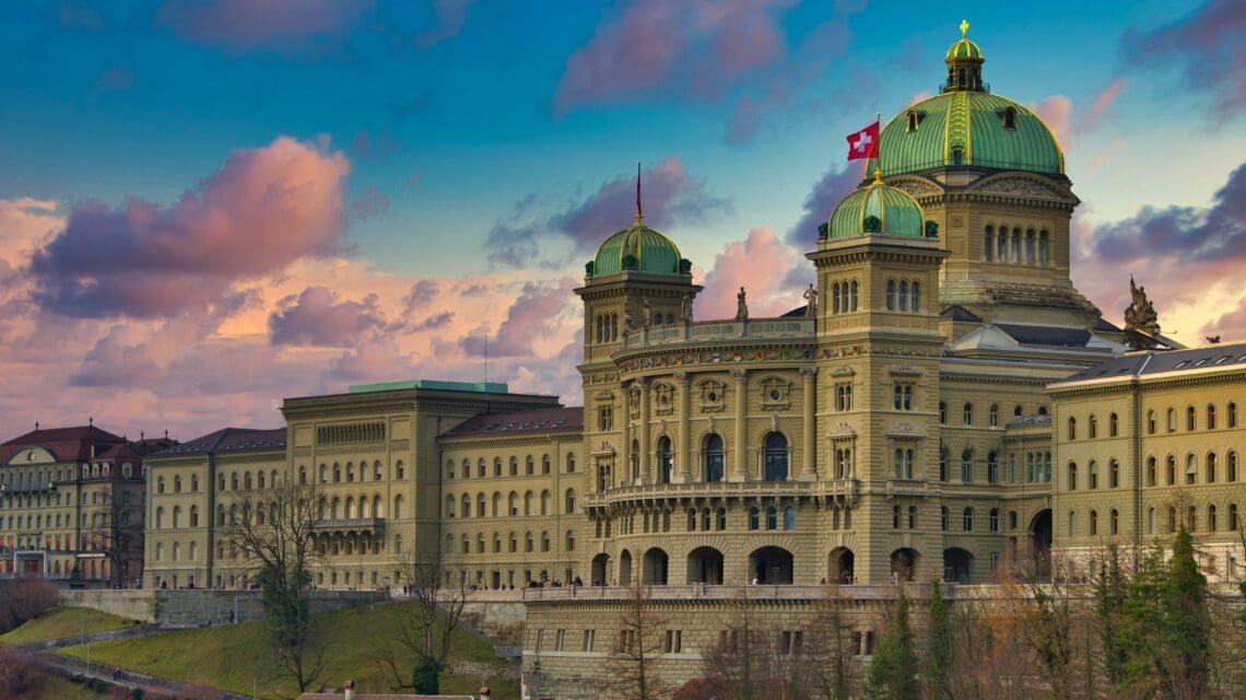 A grand, historic building with green domes and a Swiss flag, set against a vibrant sunset sky with pink clouds, evokes a serene, majestic atmosphere.