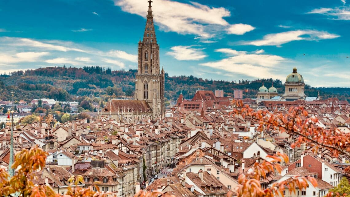 Panoramic view of a historic European city with a tall Gothic cathedral tower and domed buildings. Red roofs are framed by autumn foliage under a blue sky.