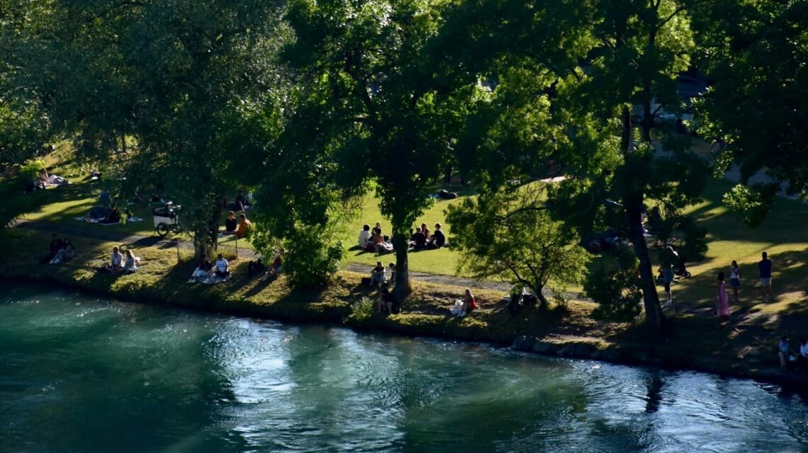 A peaceful riverside scene with people relaxing on a grassy bank beneath lush trees. In the background, charming houses are nestled against a forested hillside.