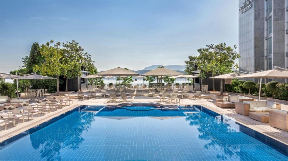 Luxurious outdoor pool area with clear blue water, surrounded by sun loungers and umbrellas. Flanked by trees and a tall building. Calm, inviting atmosphere.