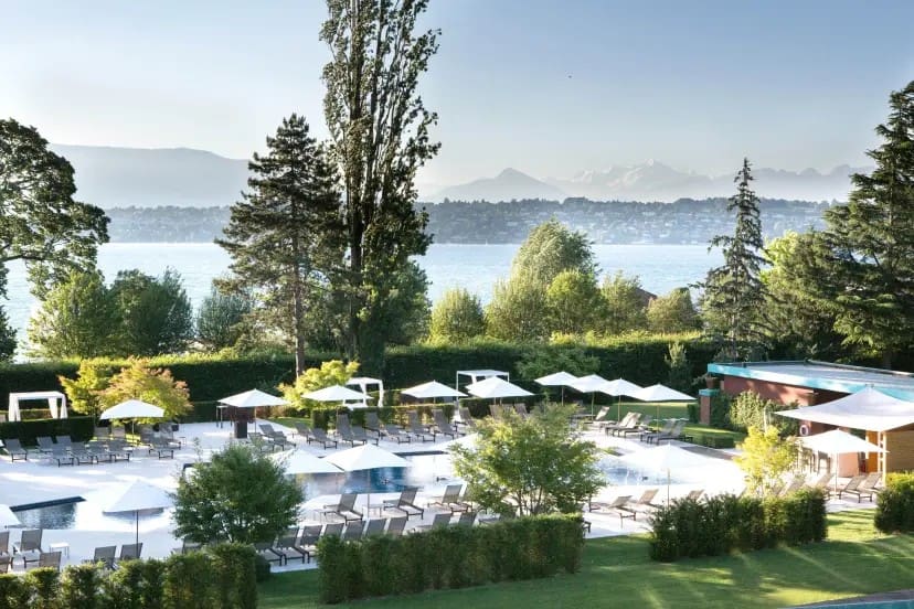 A serene outdoor pool area with white umbrellas surrounded by lush green trees, overlooking a tranquil lake and distant mountains under a clear sky.