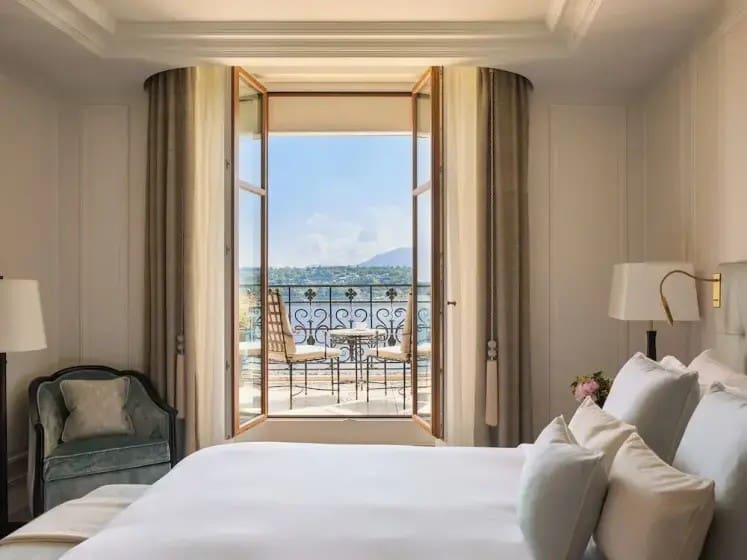 Cozy bedroom with a white bed and elegant decor, leading to a balcony with a table and chairs. The view showcases a tranquil lake and mountains.