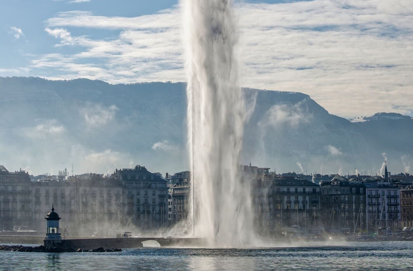 A towering fountain shoots a powerful stream of water skyward against a clear blue sky, with city buildings and distant mountains in the background.