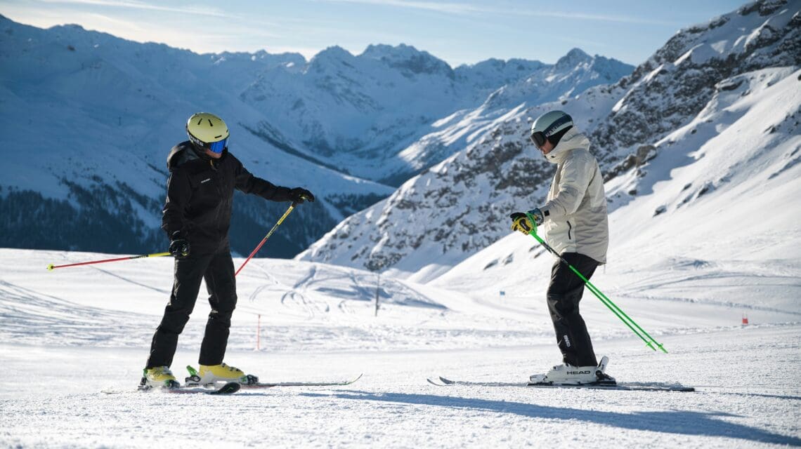 Two skiers stand on a snowy slope under a clear blue sky, surrounded by majestic mountain peaks. Both appear focused and engaged in a lesson.