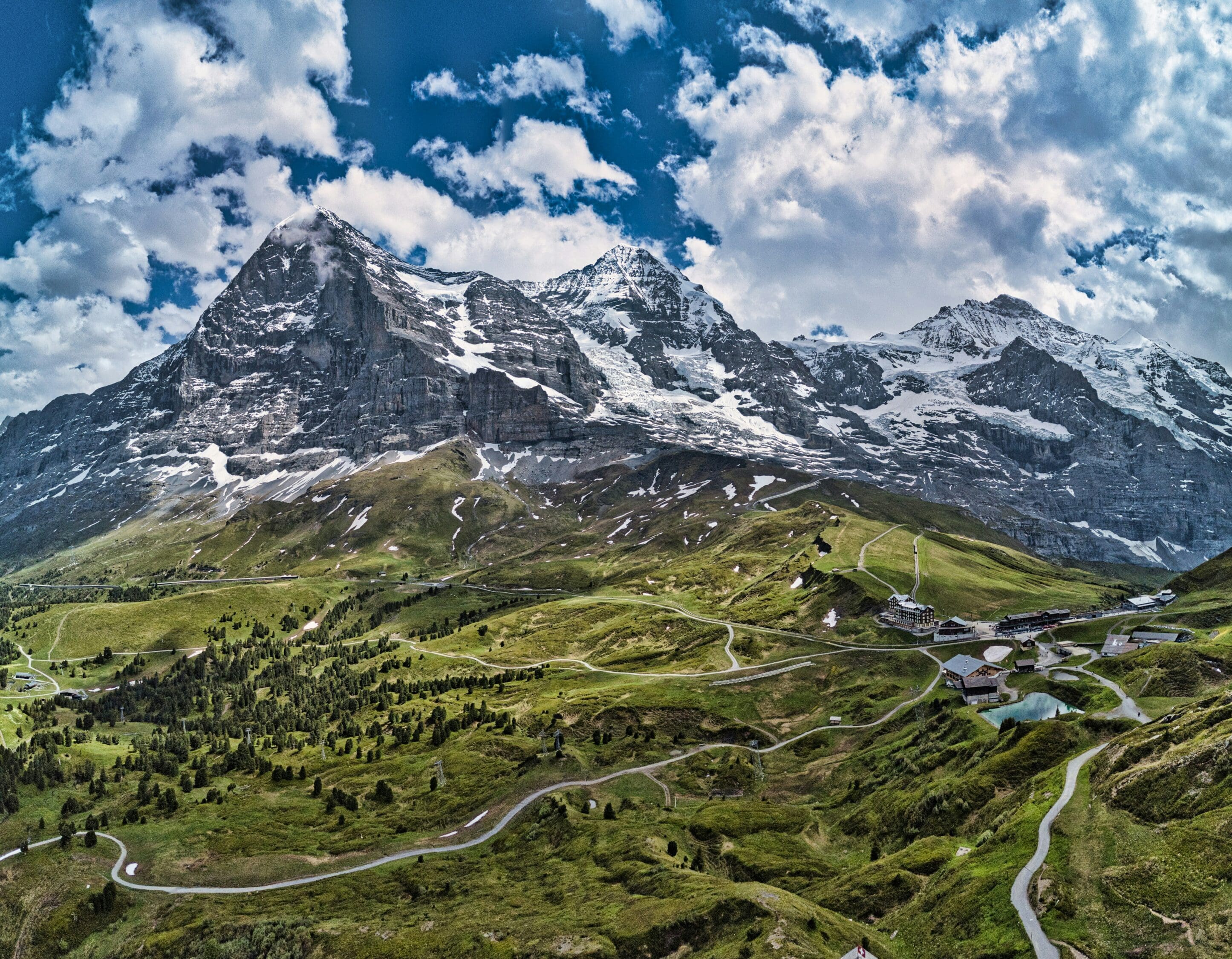 Snow-capped mountains under a partly cloudy sky, with green hills and winding roads in the foreground, creating a majestic and serene landscape.