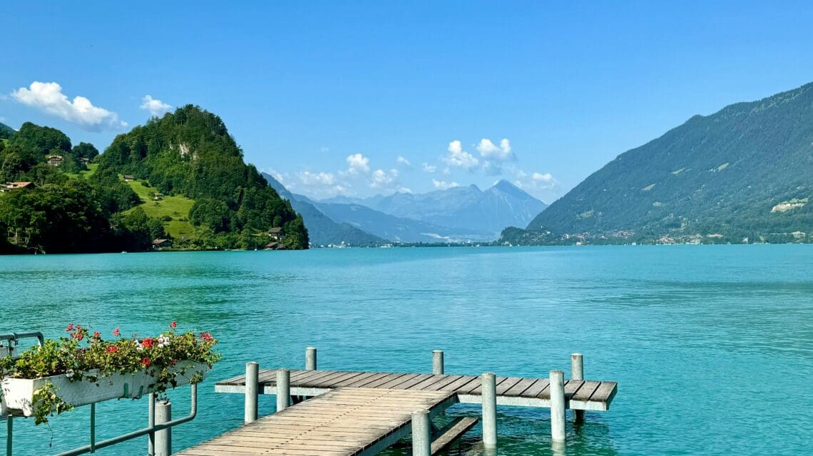 A wooden pier extends into a serene turquoise lake, surrounded by lush green hills and distant mountains under a clear blue sky. A tranquil and scenic setting.