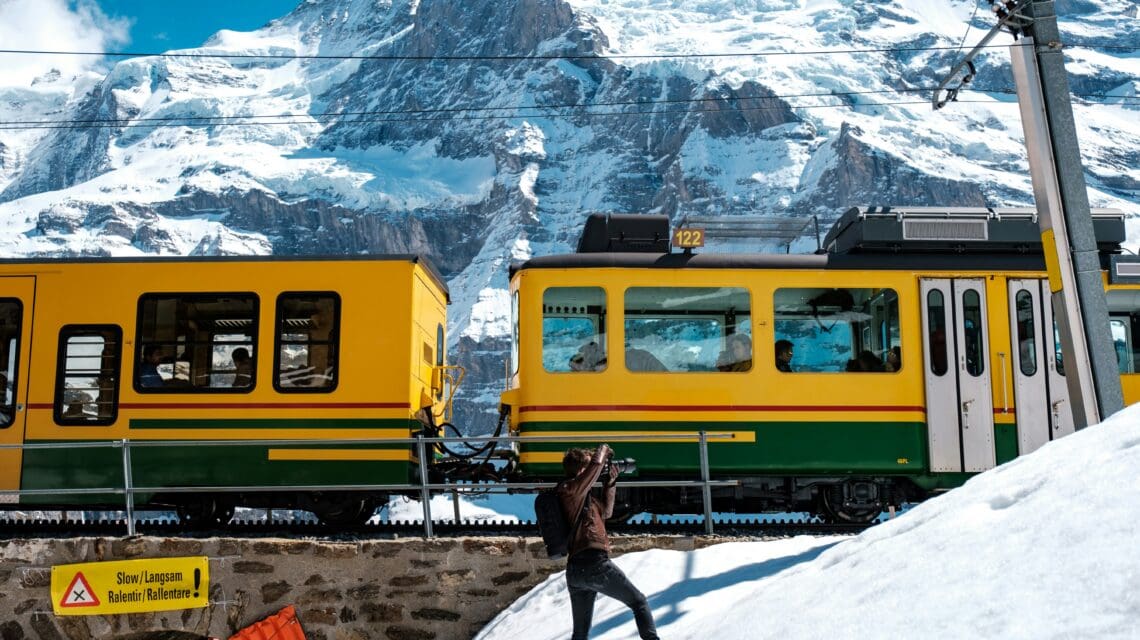 A person in a winter coat photographs a passing yellow and green train against a snow-covered mountain backdrop. The scene is bright and serene.