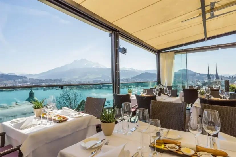 Outdoor restaurant with elegantly set tables overlooks serene lake and stunning snow-capped mountains in the distance. Sunny, inviting atmosphere.