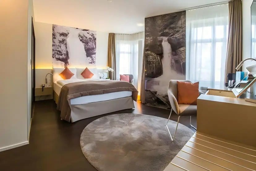 Modern hotel room with a cozy ambiance. Features a double bed with brown and white linens, waterfall mural, round rug, and seating area with soft lighting.