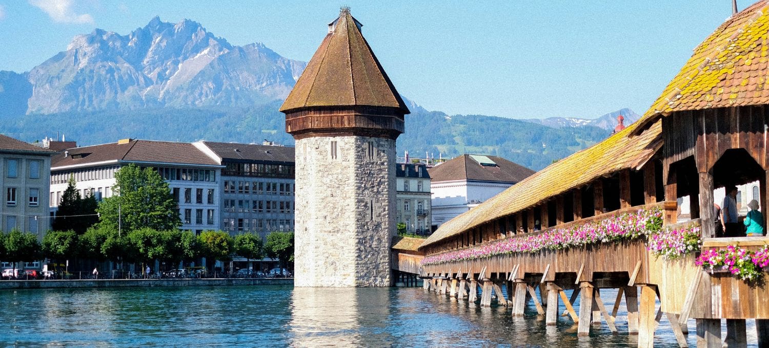A picturesque scene of Lucerne's Chapel Bridge with its stone water tower, surrounded by blue waters, flowers, and distant mountains under a clear sky.