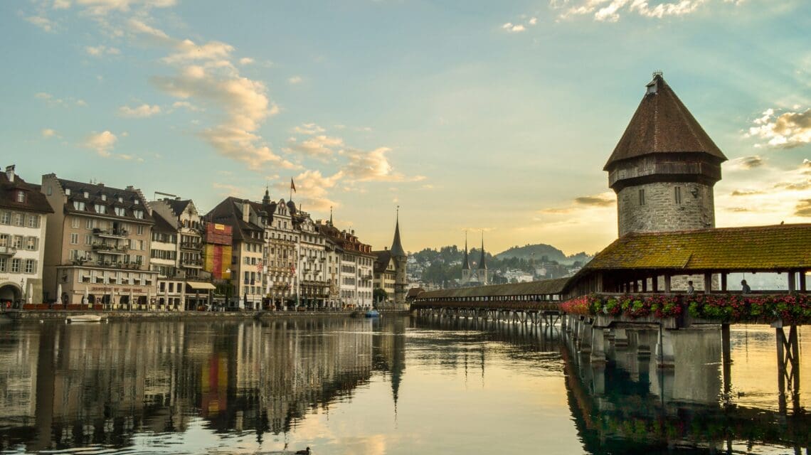Historic wooden bridge with floral decorations and water tower reflecting in a calm river. Quaint buildings and a church steeple under a soft sunset sky.