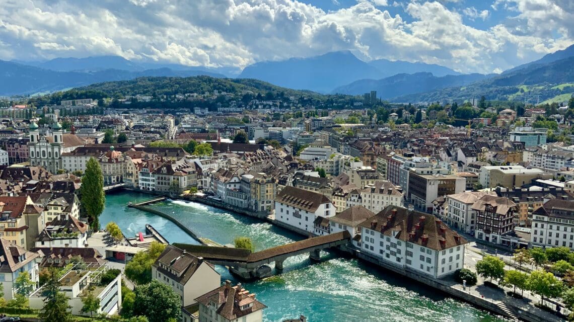 Aerial view of a picturesque European town with a river running through, stone bridge, historic buildings, hills in the distance, and a bright sky.