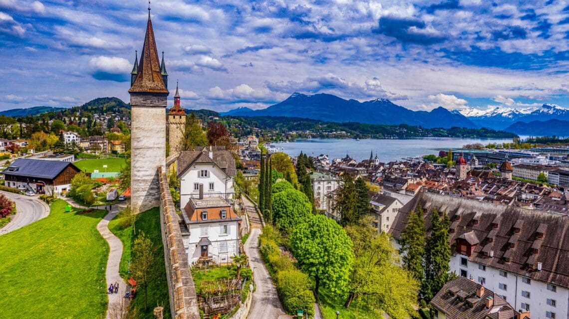 Aerial view of a picturesque town by a lake with lush greenery. A historic tower and city wall stand out, framed by distant mountains under a cloudy sky.