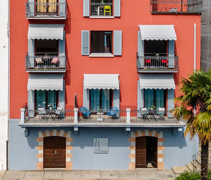 A vibrant red building with blue shutters and six balconies, each with white awnings. A palm tree adds a tropical feel against the cheerful facade.