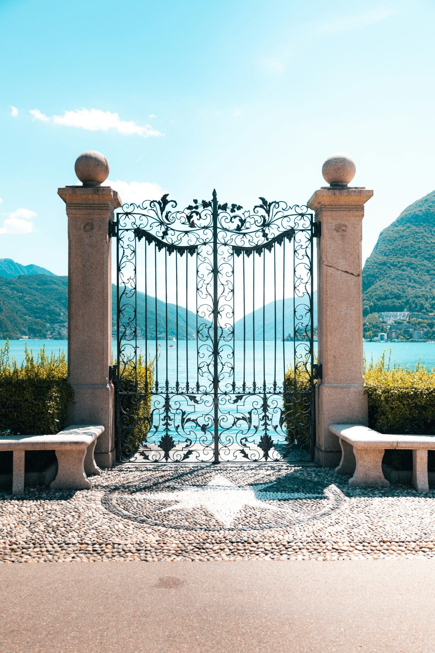 Ornate wrought iron gate flanked by stone benches, overlooking a tranquil blue lake with mountains in the background under a clear sky. Peaceful ambiance.