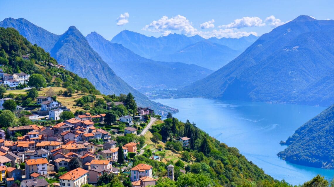 Picturesque village with orange rooftops nestled on a hillside, overlooking a serene lake and surrounded by towering green mountains under a clear blue sky.