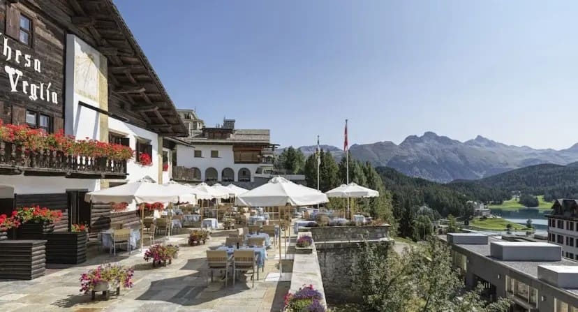 Charming outdoor restaurant with wooden architecture, red flowers, and white umbrellas, overlooking mountains and a serene lake under a clear blue sky.