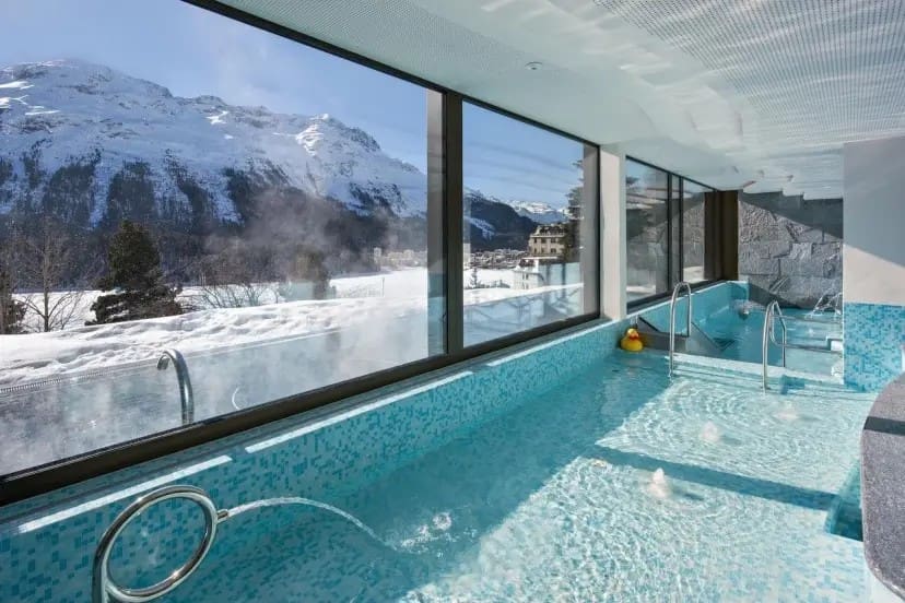 Indoor pool with blue mosaic tiles, large windows showing snowy mountains and clear sky. Steam rises from the water, creating a serene, luxurious feel.
