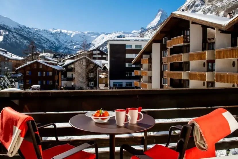 A cozy balcony with red-draped chairs and a table with mugs overlooks a snowy alpine village and the iconic Matterhorn under a clear blue sky. Relaxed ambiance.