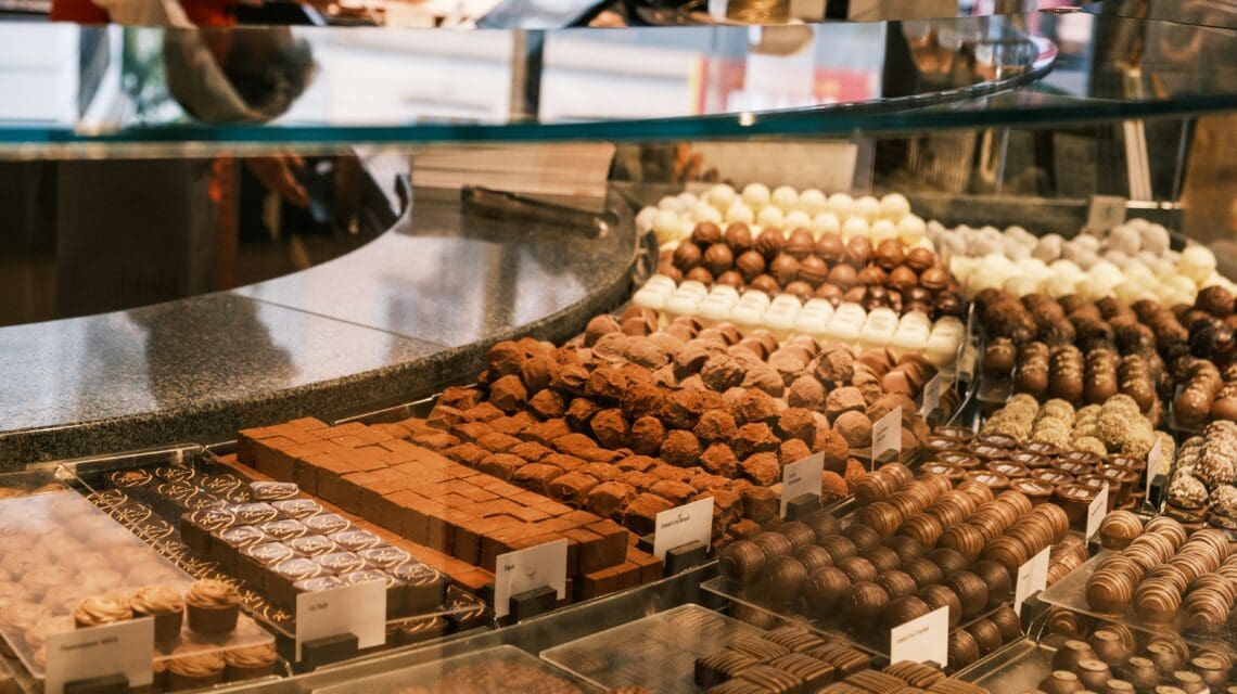 Assorted chocolates displayed in a glass case at a shop. Varieties include truffles, pralines, and squares. Warm, inviting atmosphere with rich colors.