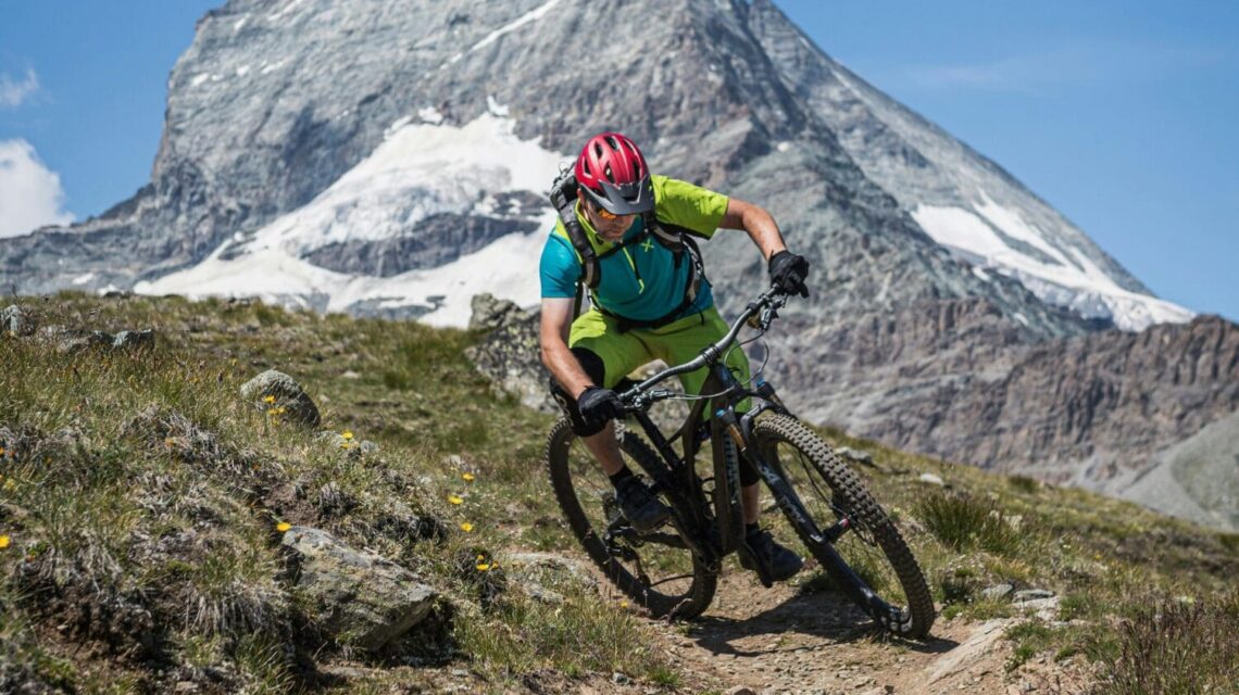 Mountain biker in green shirt and red helmet rides downhill on a rugged trail with the Matterhorn in the background under a clear blue sky.