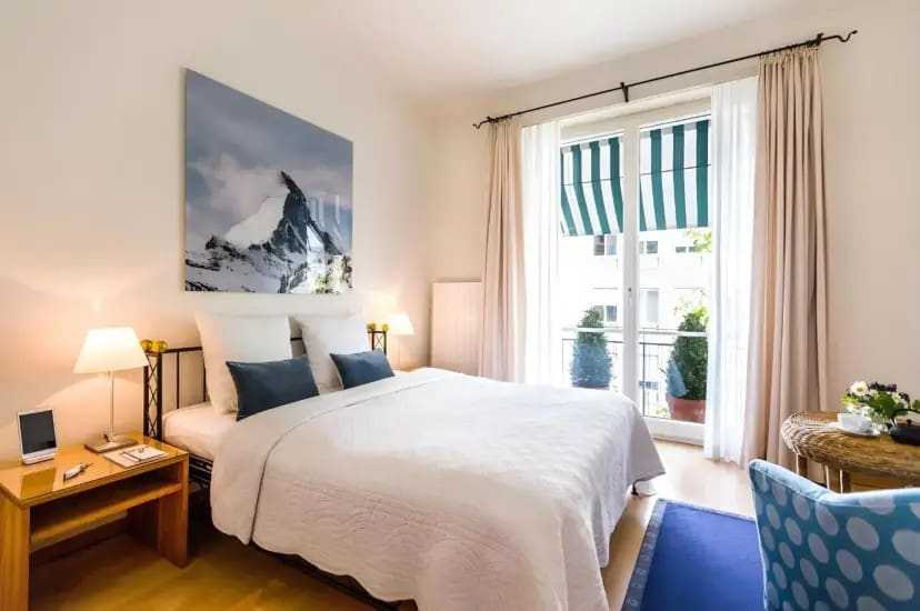 Bright, cozy bedroom with a large bed, white bedding, and blue cushions. A mountain photo hangs above. Sunlit balcony with striped awning visible.
