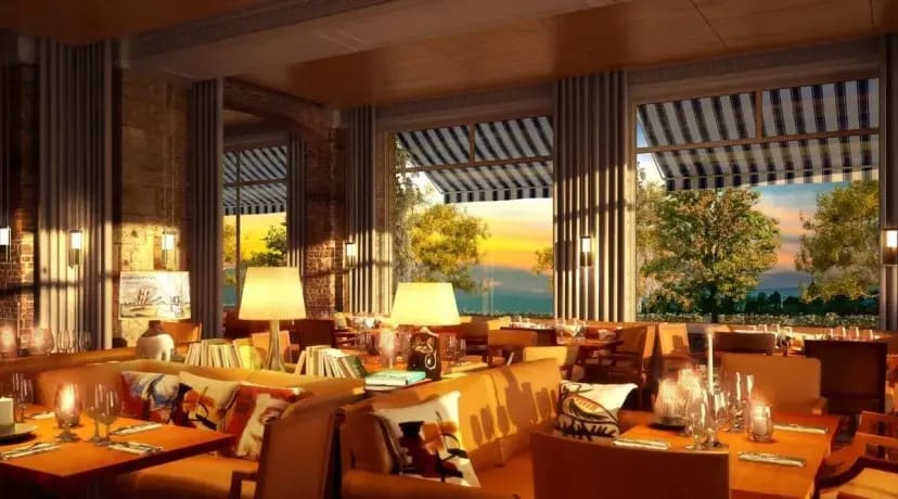 Sunlit restaurant interior with elegant wooden tables and comfy orange sofas, adorned with patterned cushions. Large windows reveal a vibrant sunset sky.
