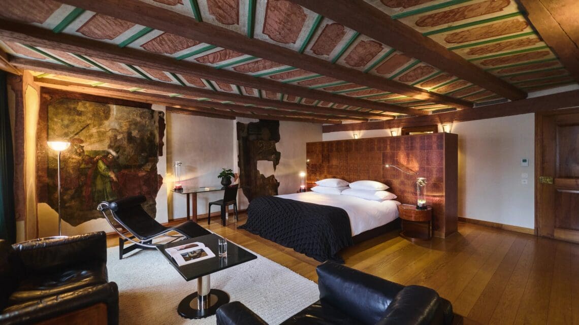 luxurious hotel room with wooden ceiling, large bed with dark bedding, lounge chairs, modern art, and warm lighting; cozy and inviting ambiance.