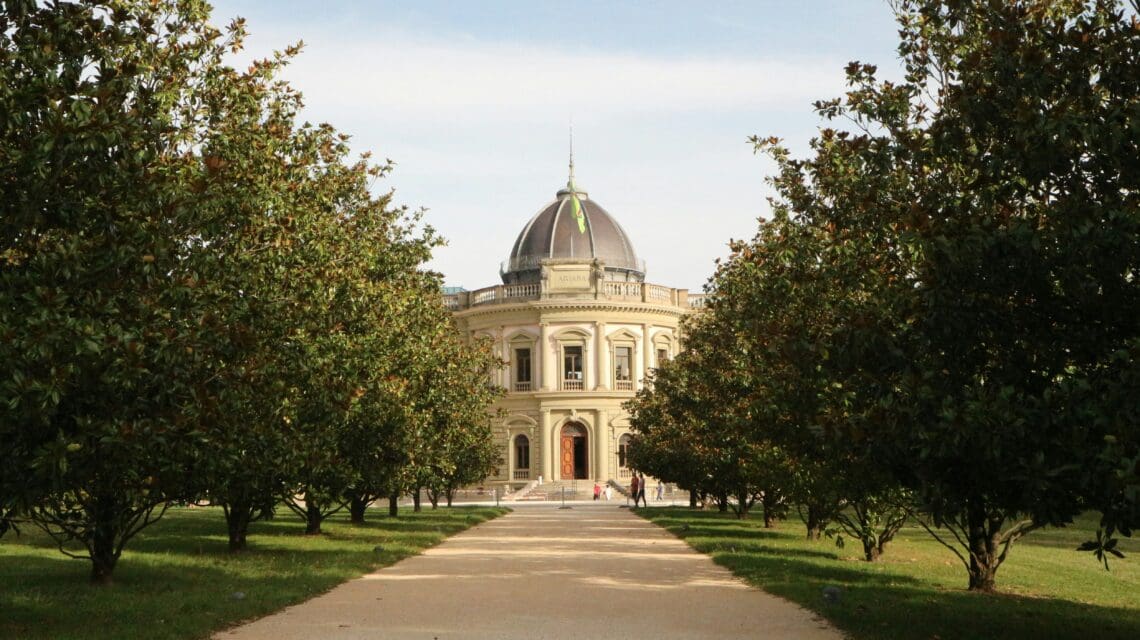 A grand rotunda with a dome, framed by symmetrical rows of lush green trees on either side of a wide path. The scene feels serene and majestic.