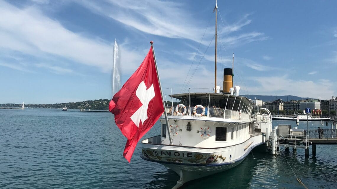 A vintage boat with a Swiss flag is moored on a calm lake, with a distant fountain and mountains under a blue sky. The scene is peaceful and picturesque.