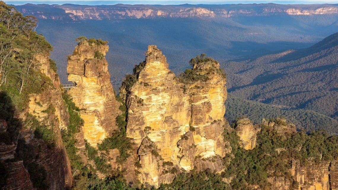 A scenic view of the Three Sisters rock formation in the Blue Mountains, Australia. Sunlit sandstone cliffs rise amidst lush green forests under a clear blue sky.