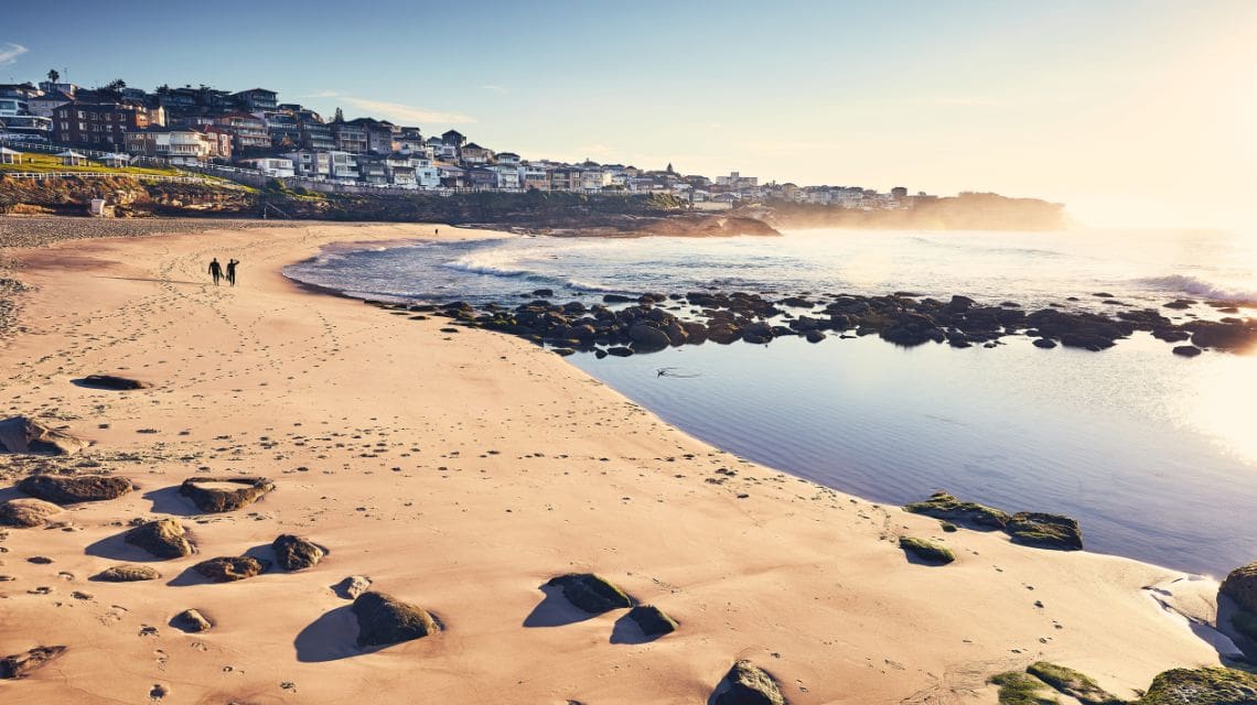 Coastal view at sunrise with sandy beach, rocks, and gentle waves. Two figures walk along the shore. Houses are visible in the background, enveloped in mist.