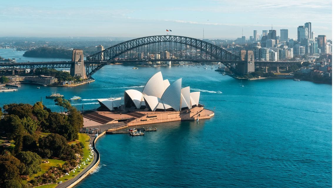 Aerial view of Sydney Opera House with its iconic white sails by blue waters. The Sydney Harbour Bridge and city skyline form the backdrop under a clear sky.