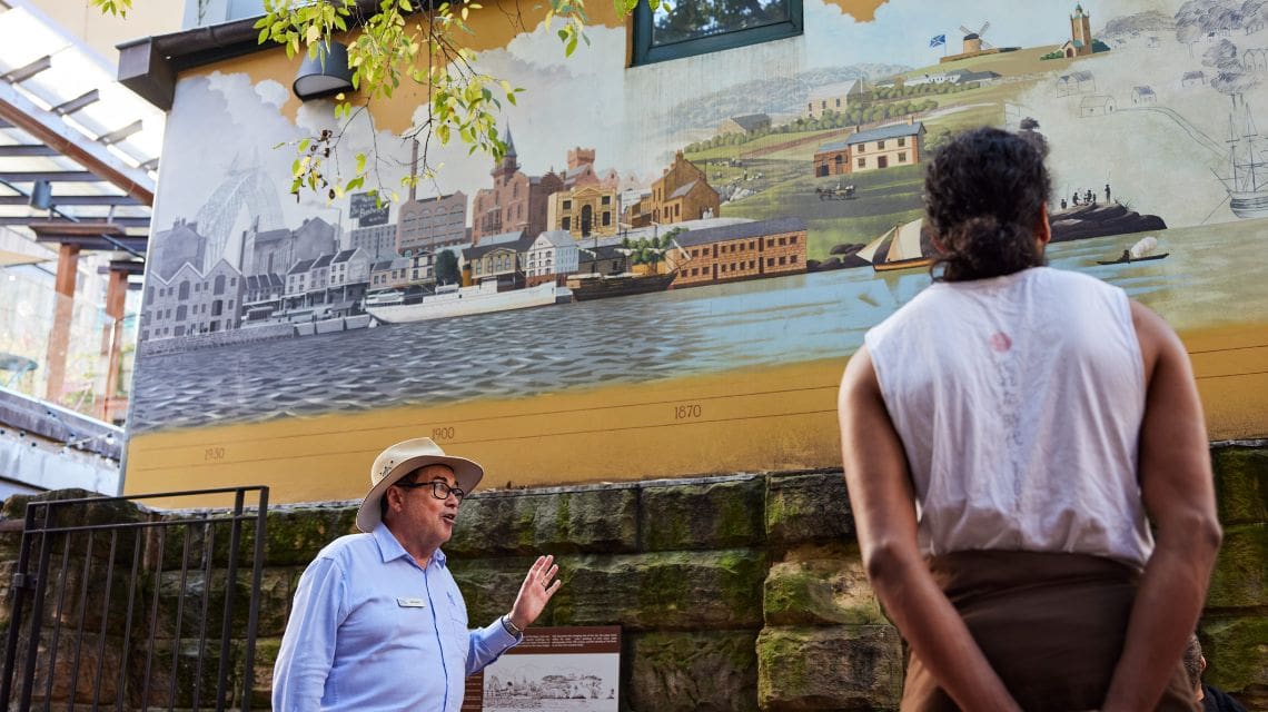 A guide gestures while describing a historical mural depicting a waterfront town scene. A visitor stands listening, conveying an educational atmosphere.