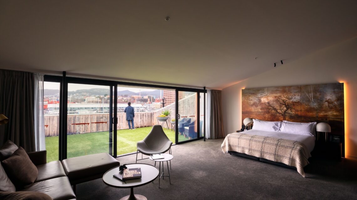 Modern hotel room with large windows opening to a balcony with a city and harbor view. Cozy decor, warm lighting, and a person gazes outside.