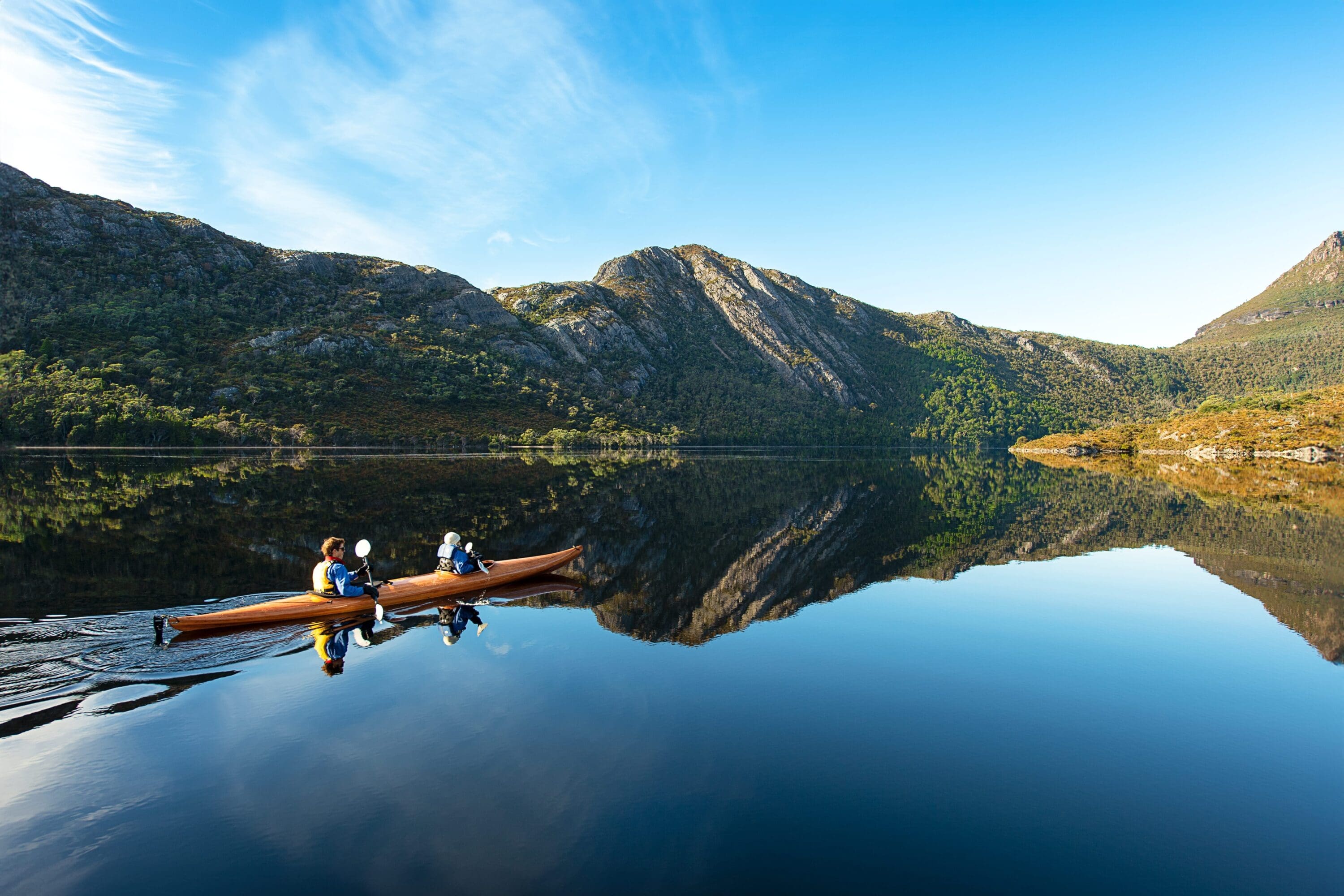 Two people canoe on a serene lake, surrounded by rugged mountains reflecting in the calm water, under a clear blue sky, creating a peaceful scene.