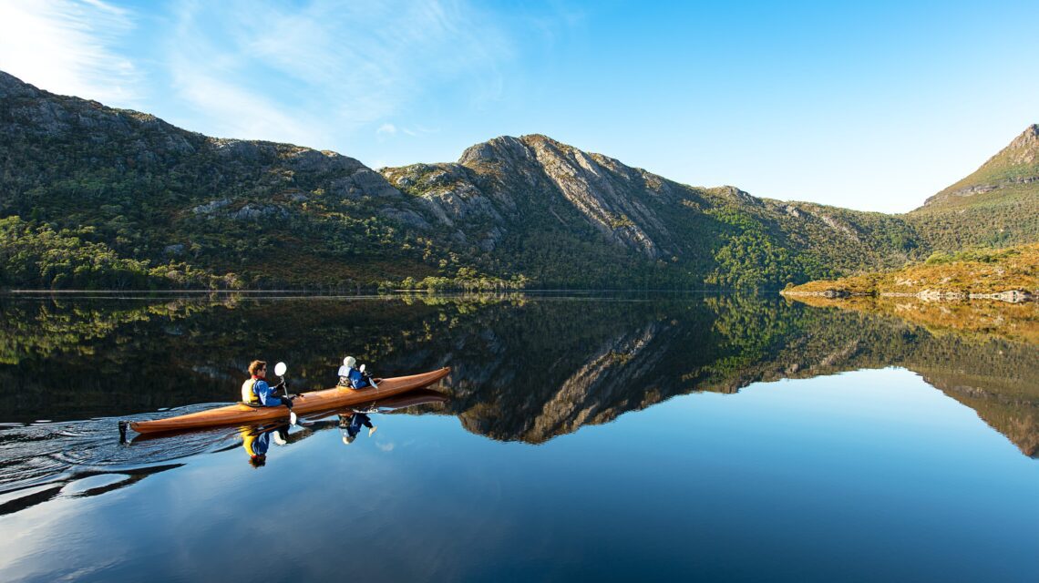 Two people kayak on a calm lake, surrounded by mountains reflecting in the water under a clear blue sky, evoking tranquility and adventure.