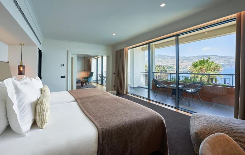Luxurious hotel room with a large bed and soft lighting. A spacious balcony offers stunning ocean and mountain views, creating a tranquil atmosphere.