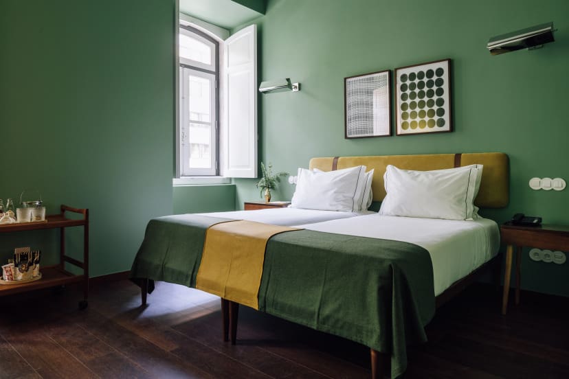 A cozy bedroom with green walls, twin beds draped with green and yellow blankets, white pillows, and minimal decor. Soft light from an open window creates a calm ambiance.