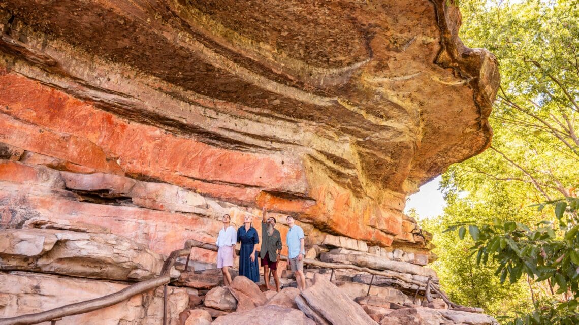 Four people stand under a massive, layered rock formation with red and orange hues. Bright greenery surrounds the area, conveying a sense of adventure.