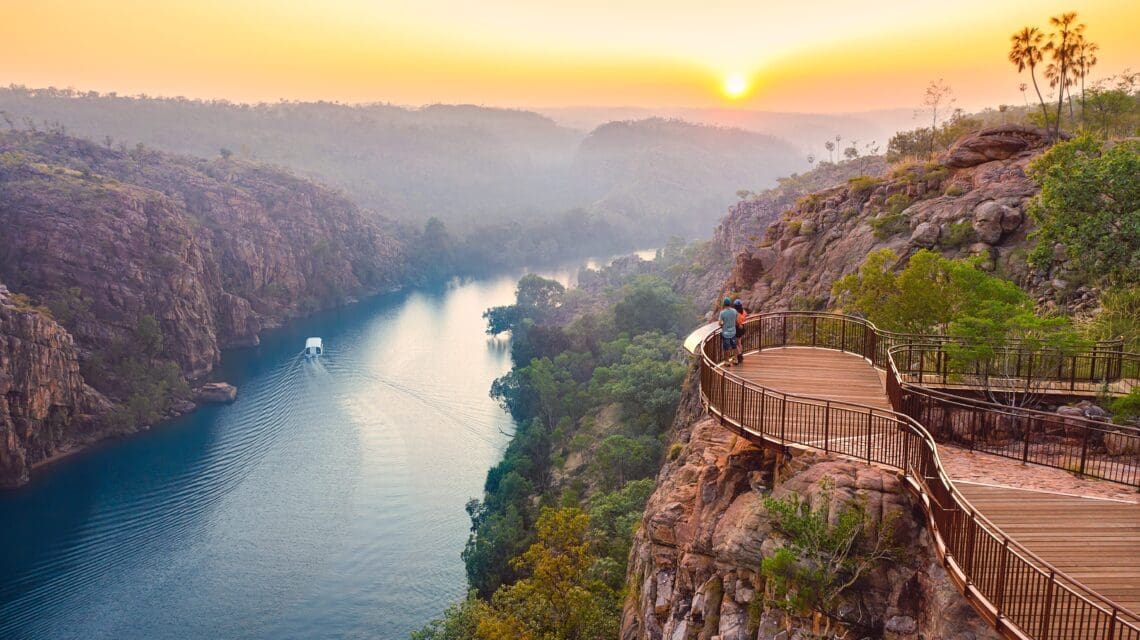 Sunset view from an elevated wooden walkway overlooking a serene river winding through lush green cliffs. A tranquil boat glides below.