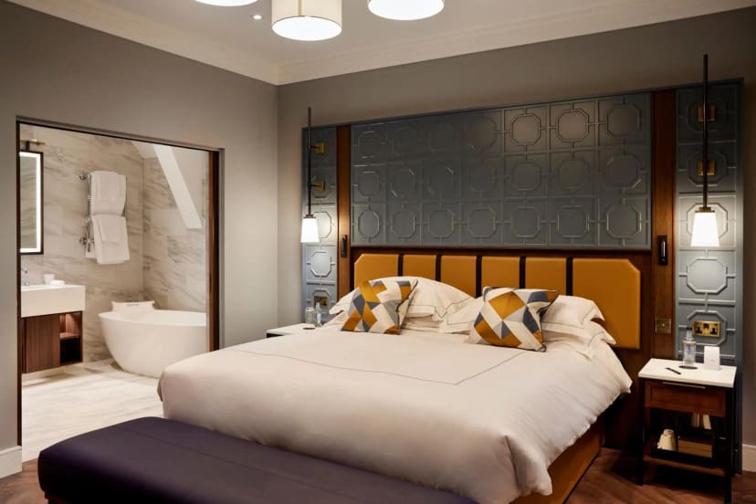 Stylish hotel room with a plush double bed, geometric pillows, ornate headboard, and soft lighting. Bathroom with a modern tub is visible, creating a luxurious ambiance.