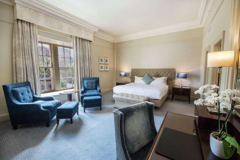Elegant hotel room with a king-sized bed, blue armchairs, and a window overlooking greenery. Soft lighting and neutral tones create a calm atmosphere.